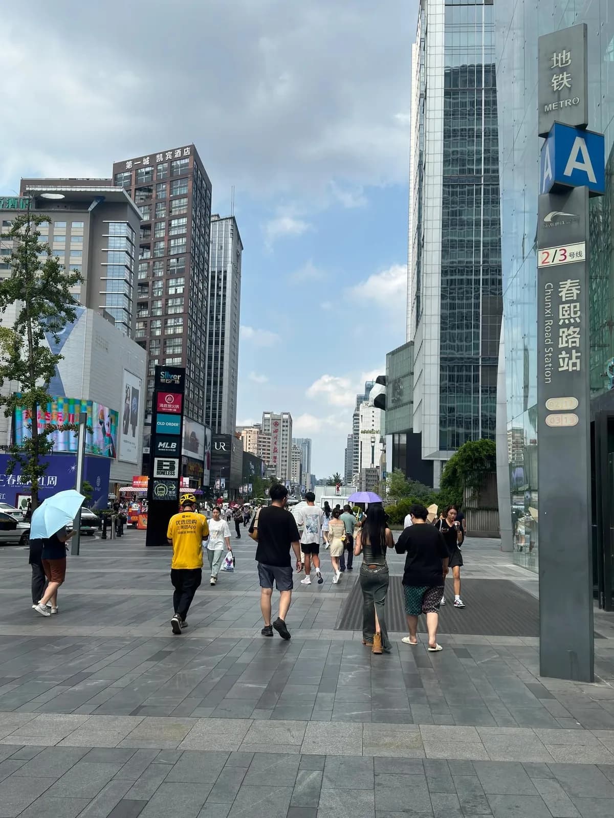 Pedestrians on Chunxi Road beside a metro entrance sign, with shops and tall buildings along the street.