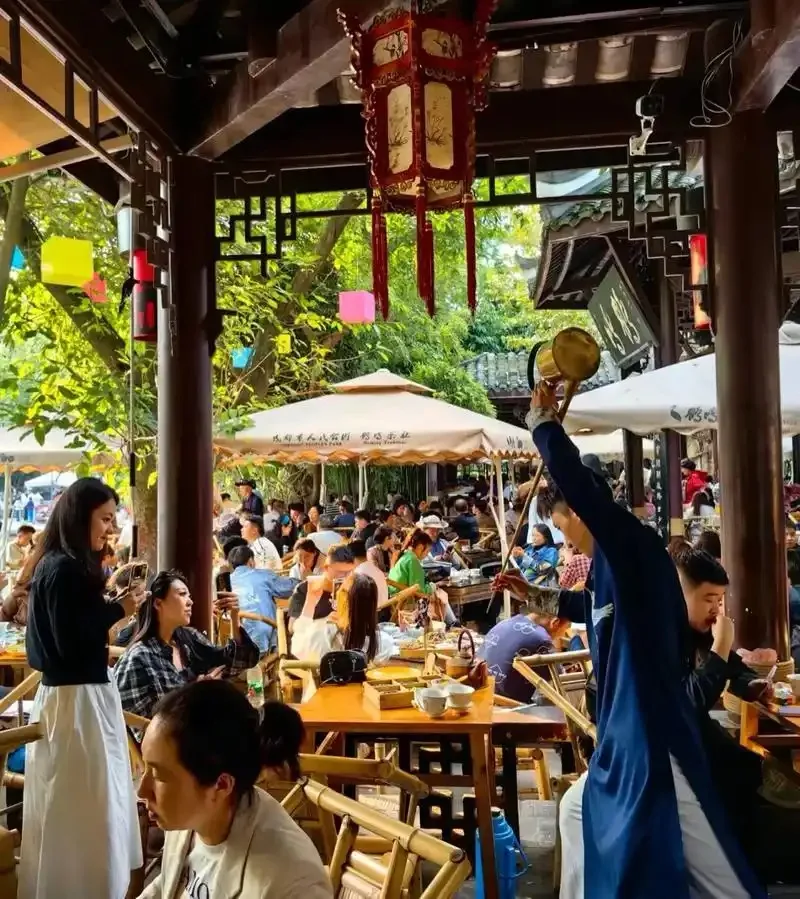 A tea server raises a long-spouted kettle beside tables and bamboo chairs in a busy Chengdu teahouse.