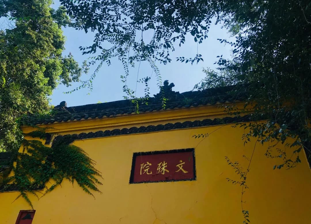 A yellow wall with the Chinese name of Wenshu Monastery beneath overhanging green branches.