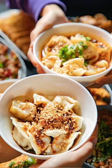 Two bowls of dumplings with red oil and chopped toppings, held above a dining table.