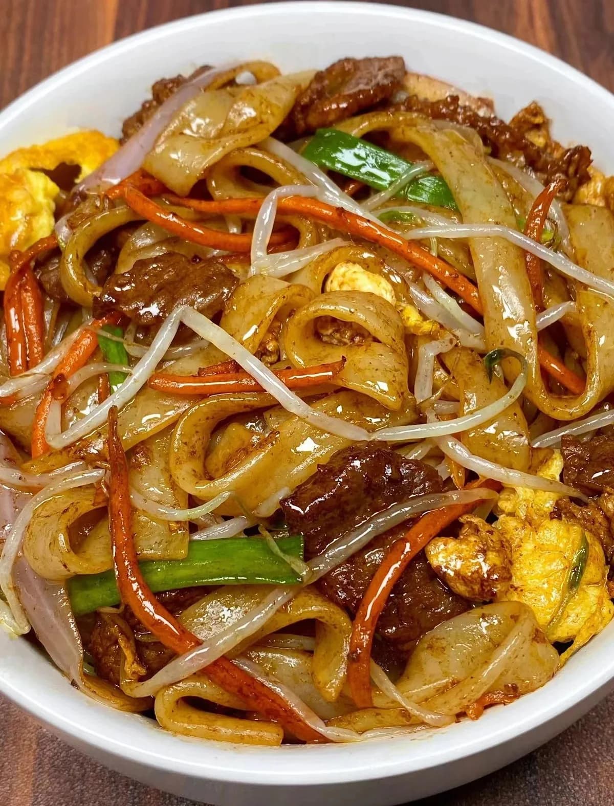 Wide rice noodles with beef, bean sprouts, vegetables and pieces of egg on a white plate.