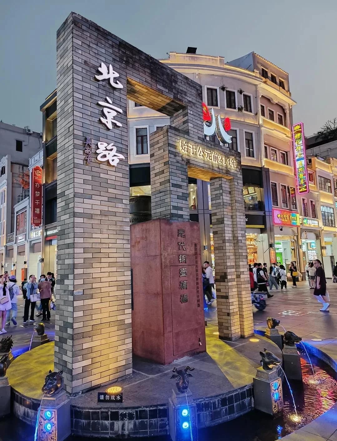 An illuminated Beijing Road monument surrounded by shopfronts and pedestrians.