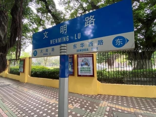 A blue Wenming Road sign beside a yellow wall and a shaded pavement.