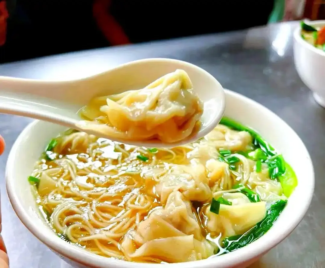 A spoon lifting a wonton above a bowl of thin noodles, broth and green vegetables.