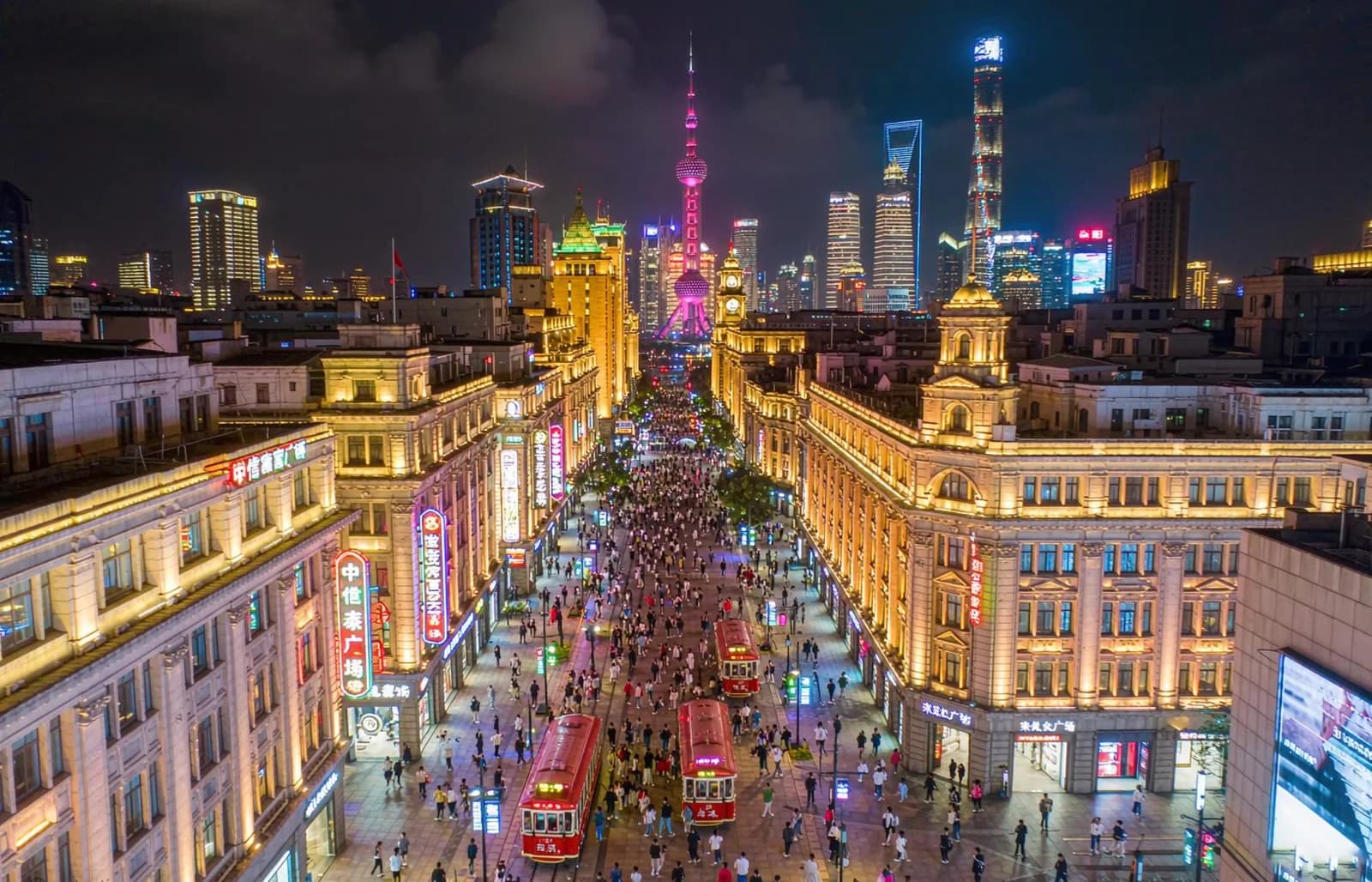 Crowds and red sightseeing vehicles on illuminated Nanjing Road, with Pudong towers in the distance.