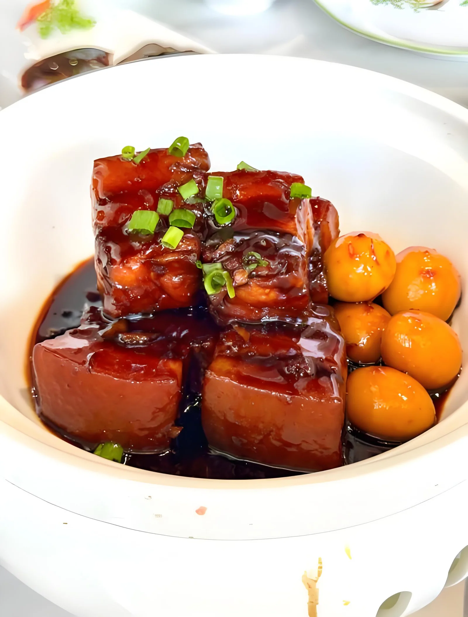 Glossy red-braised pork belly topped with scallions, with small eggs in a white bowl.