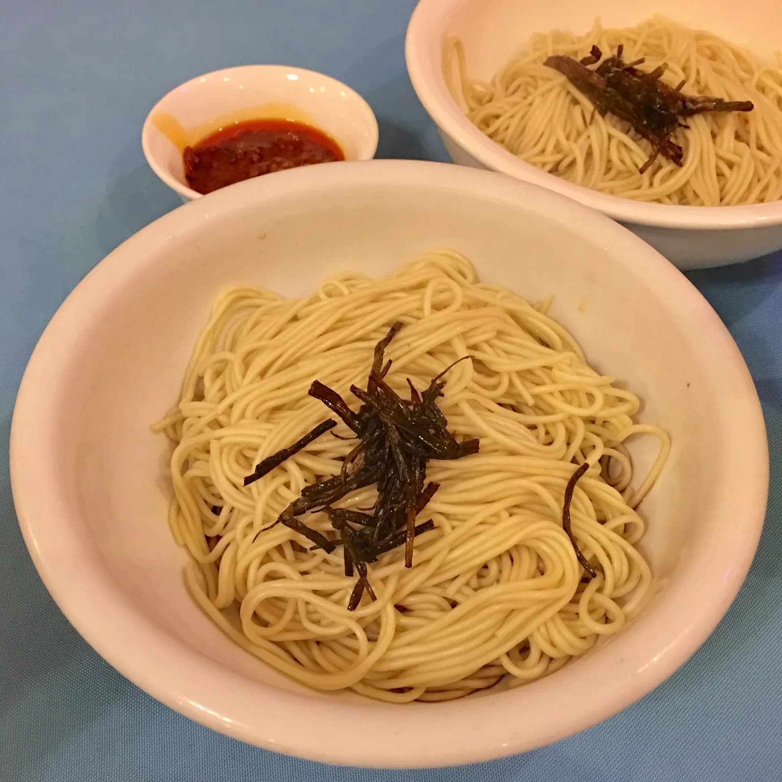 Two bowls of noodles topped with fried scallions, with a small dish of sauce.