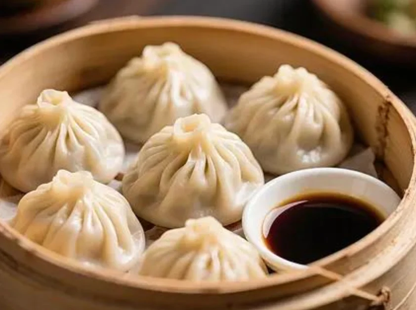 Pleated soup dumplings in a bamboo steamer beside a small dish of vinegar.