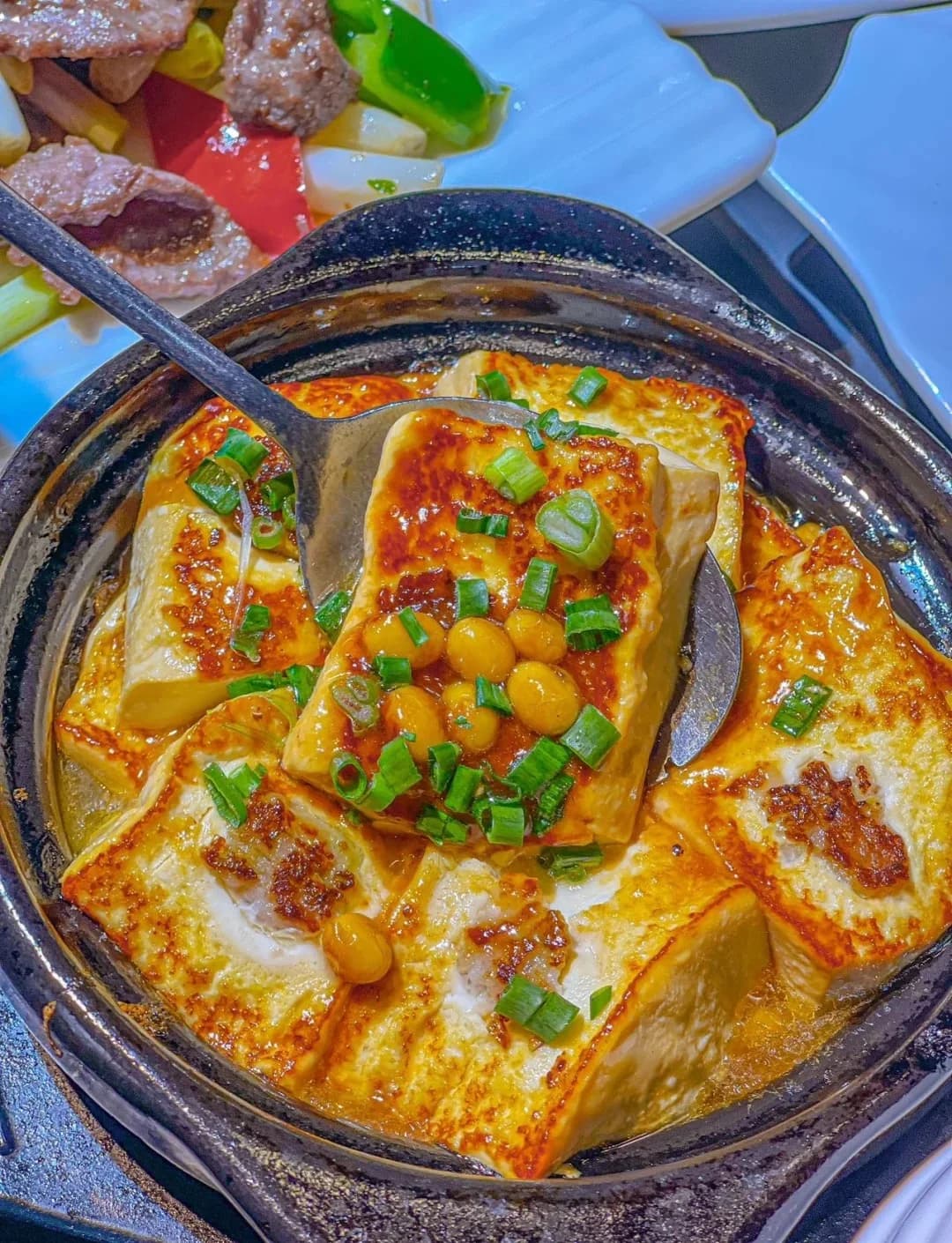 Browned tofu pieces in sauce in a dark serving pot, with a spoon lifting one piece.