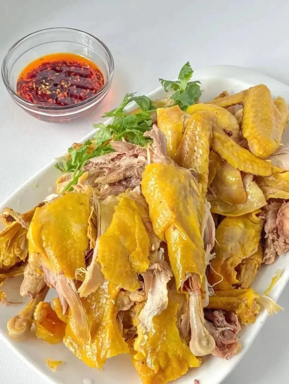 Pieces of yellow-skinned chicken on a white platter beside a small bowl of red dipping sauce.