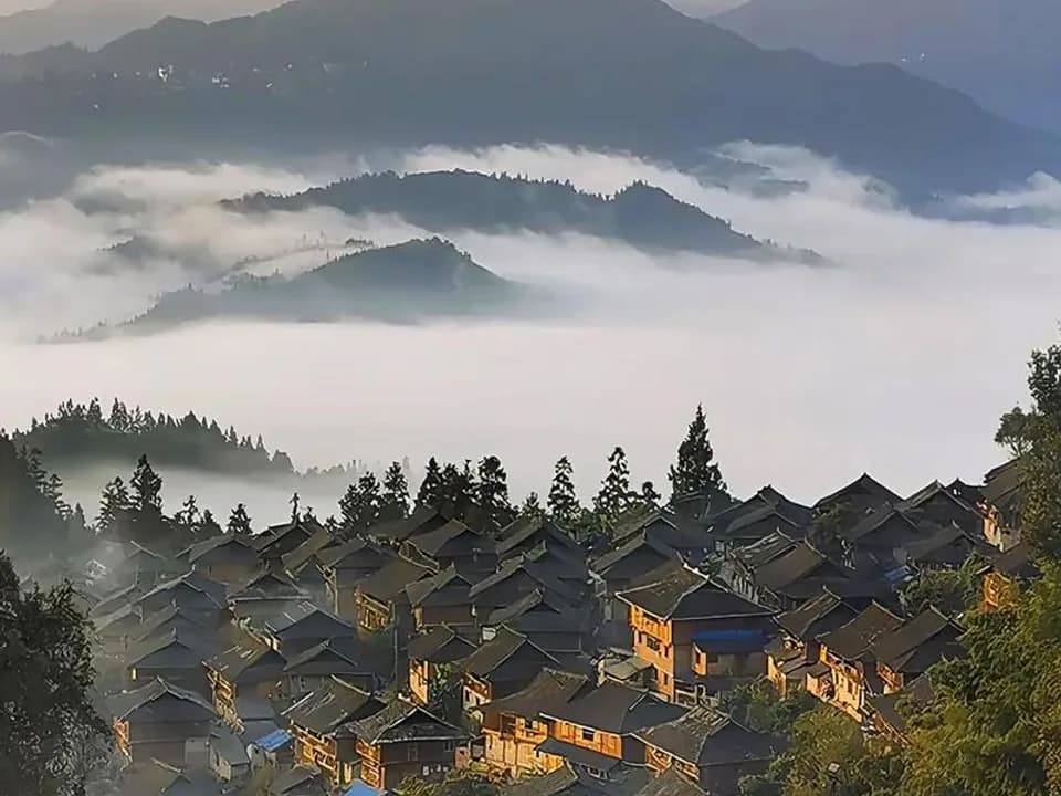 Dark-roofed mountain village below layered forested ridges and a sea of morning cloud in southeast Guizhou