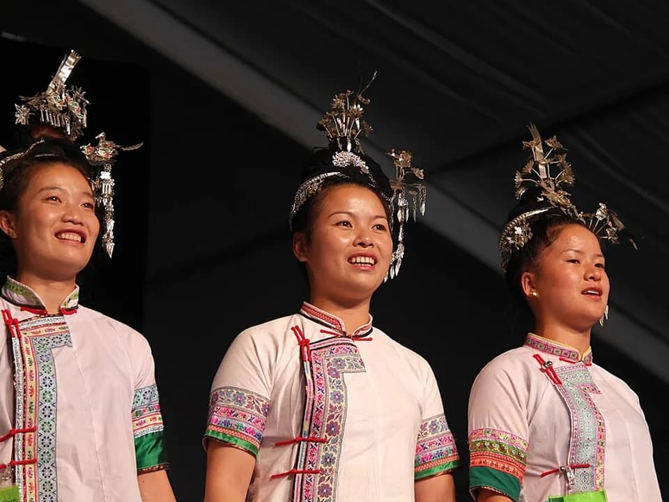 Three Dong women in embroidered clothing and silver headdresses singing together on a dark festival stage