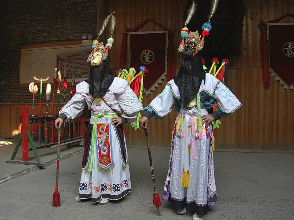 Two Tunpu Dixi performers wearing tall carved masks, ceremonial costumes and holding staffs in a Guizhou village