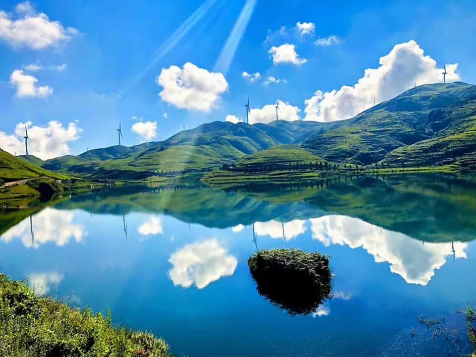 Green rolling plateau, wind turbines and cloud reflections in a blue lake at Wumeng Grassland in Panzhou